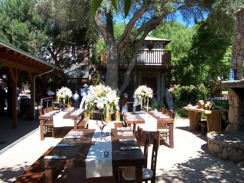 long wood tables with white table runner set up under oak tree near outdoor fireplace
